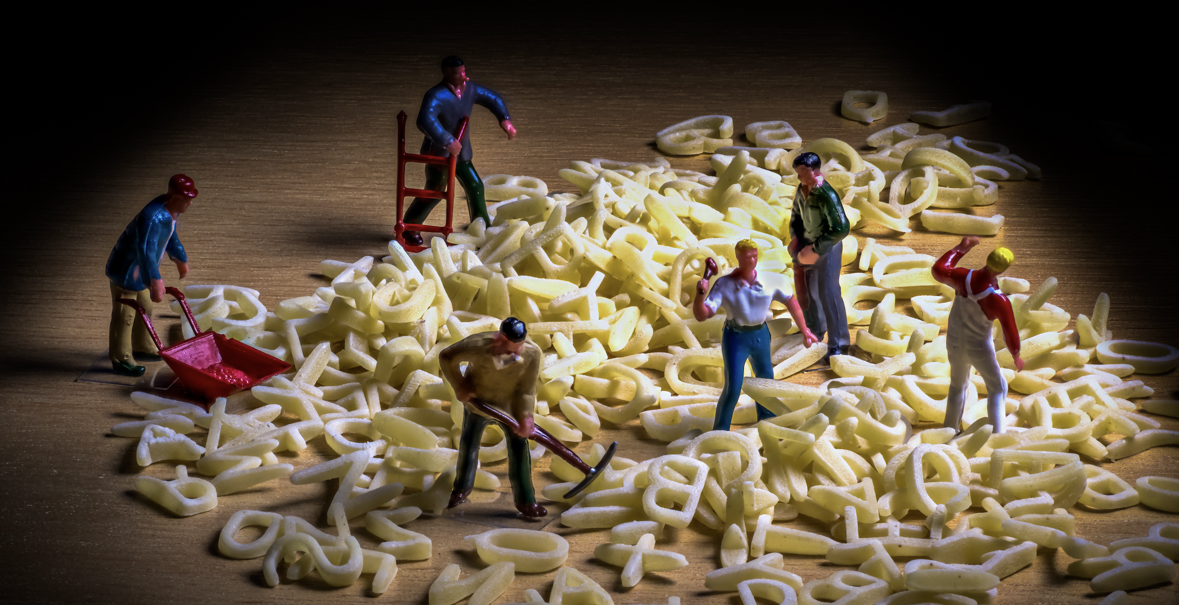 A photo of miniature toy figures depicting laborers with picks and wheelbarrows digging in a mound of alphabet-soup letters.
