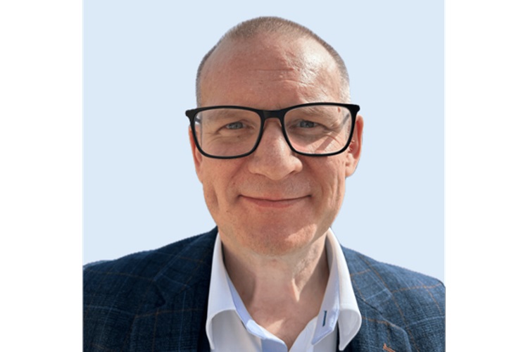 Image of a White man with close-cropped hair and large frames, wearing a gray plaid jacket.