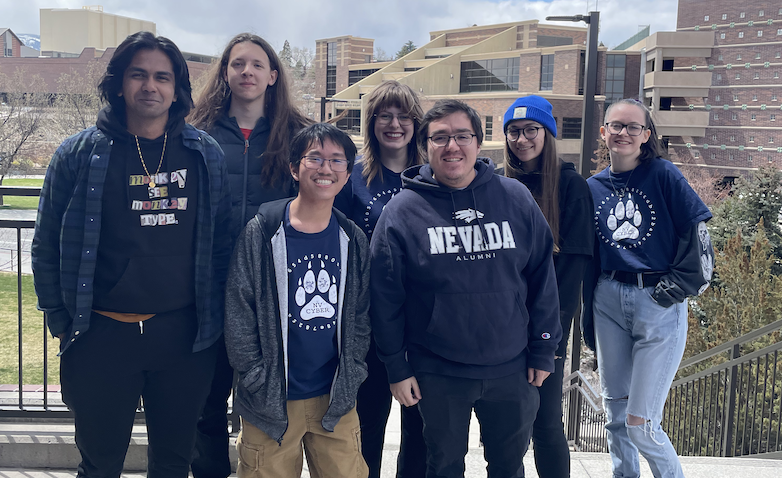 Photo of 7 people posing for a group photo on an outdoor promenade with a park and college campus buildings in the background.