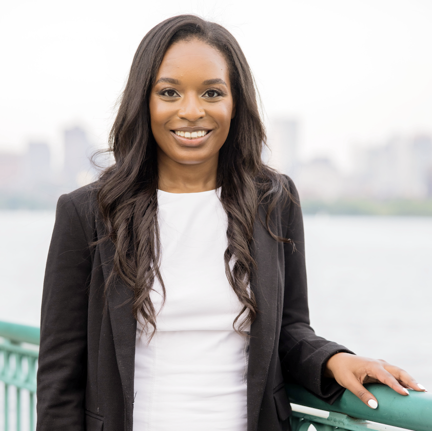 Photo of a Black woman with long straight hair in a white tee and black jacket, standing at the railing of a city waterway.