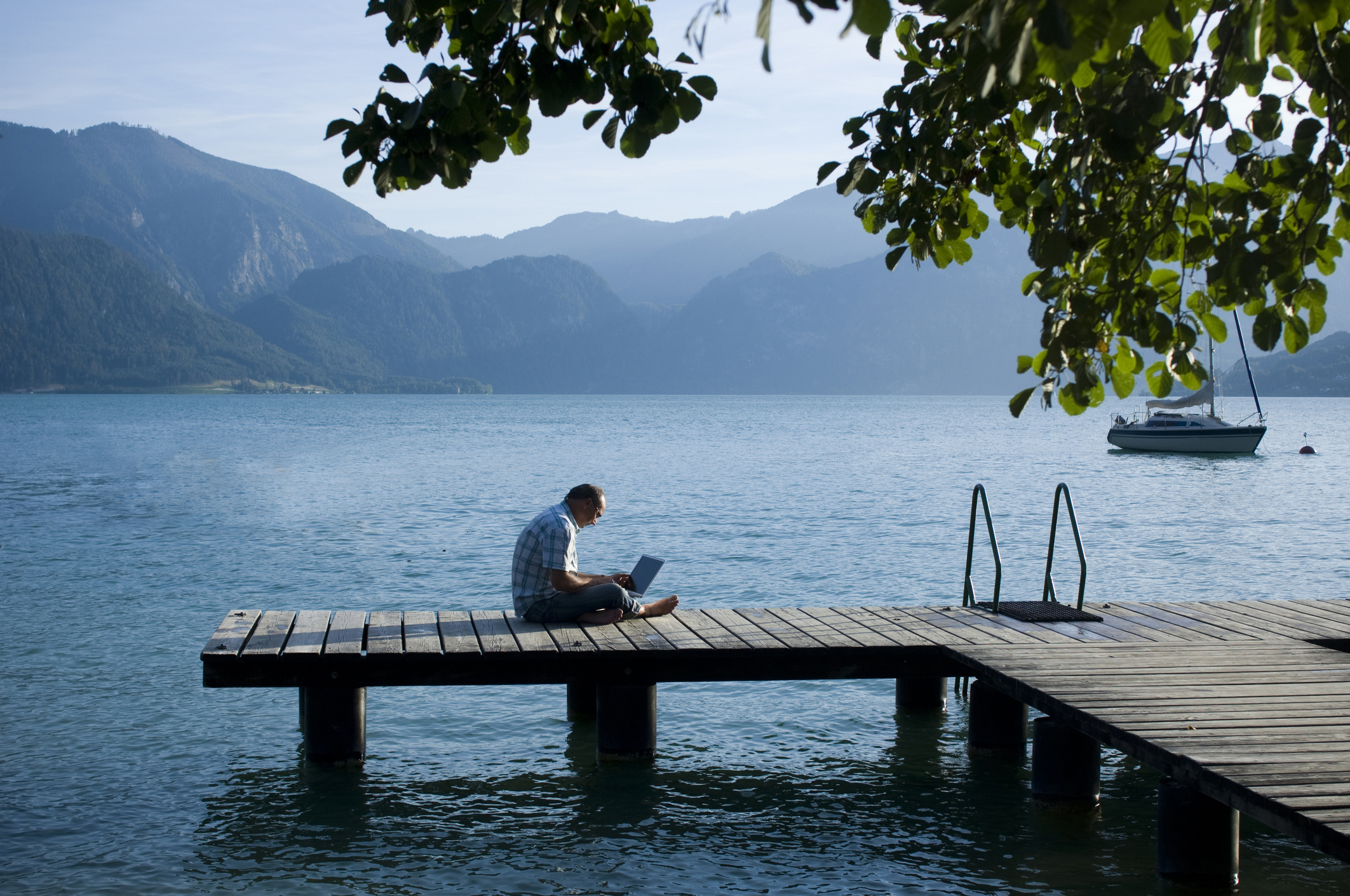 Photo of a man sitting cross-legged on a dock by a lake with a laptop in his lap.