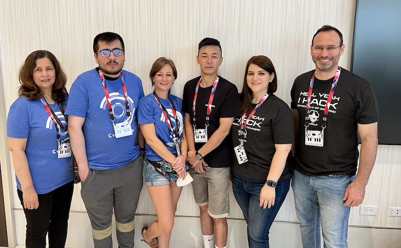 Photo of 6 people wearing matching t-shirts and convention lanyards posing for a photo in a convention hall.