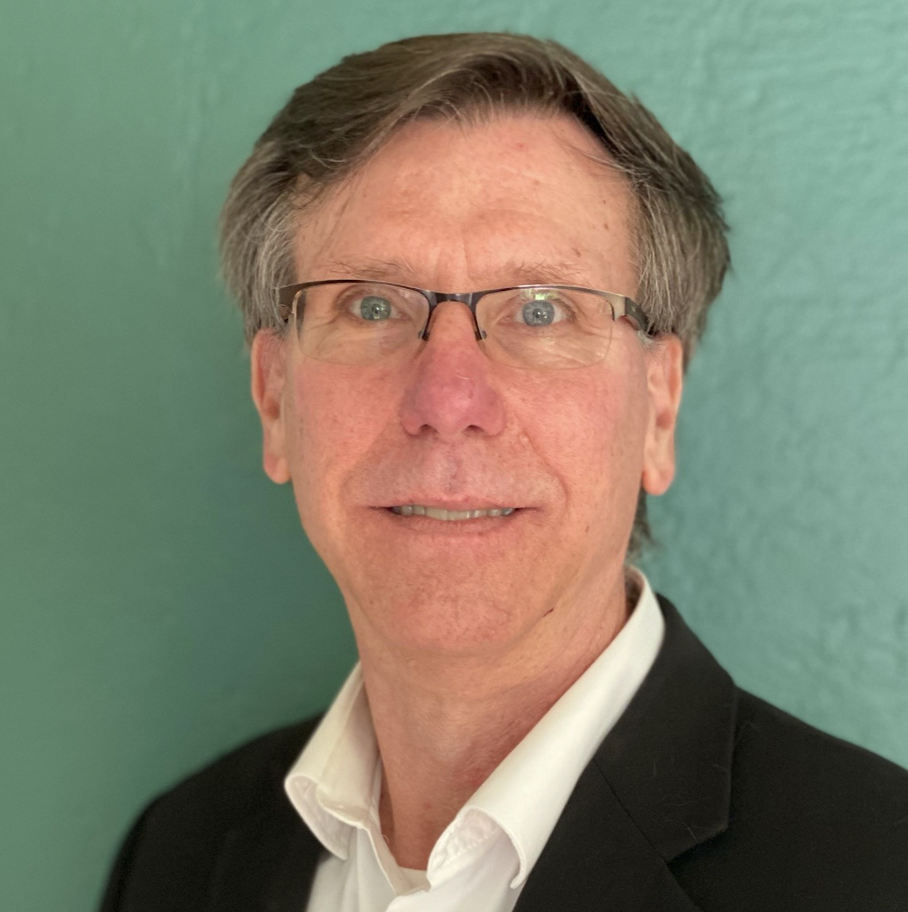 Photo of a middle-aged White man with brown hair parted on the side and glasses, wearing a black jacket and white shirt.