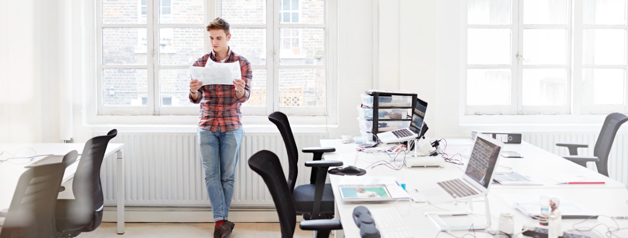 Person in flannel shirt examining papers while standing by the window in a bright white office.